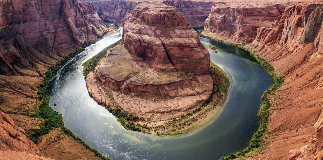 image shows horseshoe bend in the Colorado river. the water is a deep blue/green colour. there is a donut shape of water with a huge red rock in the middle. there are red cliffs surrounding all sides.