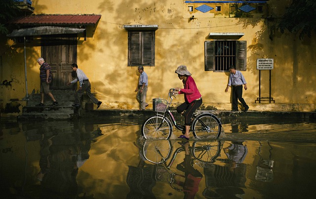 a woman in a red jacket cycles through flood waters with a house and people in the background.