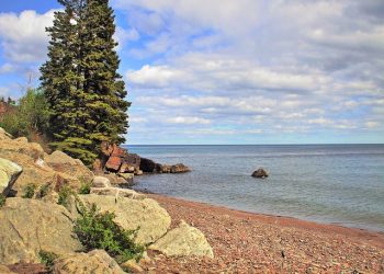 The image shows a bank of Lake Superior, in Canada. on the left is a tree and a rock embankment. On the right is the lake stretching out towards the sky. It is a blue and slightly cloudy sky.