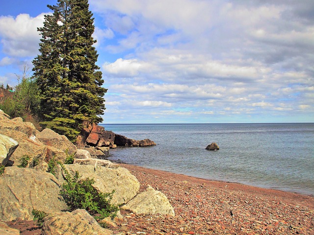 The image shows a bank of Lake Superior, in Canada. on the left is a tree and a rock embankment. On the right is the lake stretching out towards the sky. It is a blue and slightly cloudy sky.